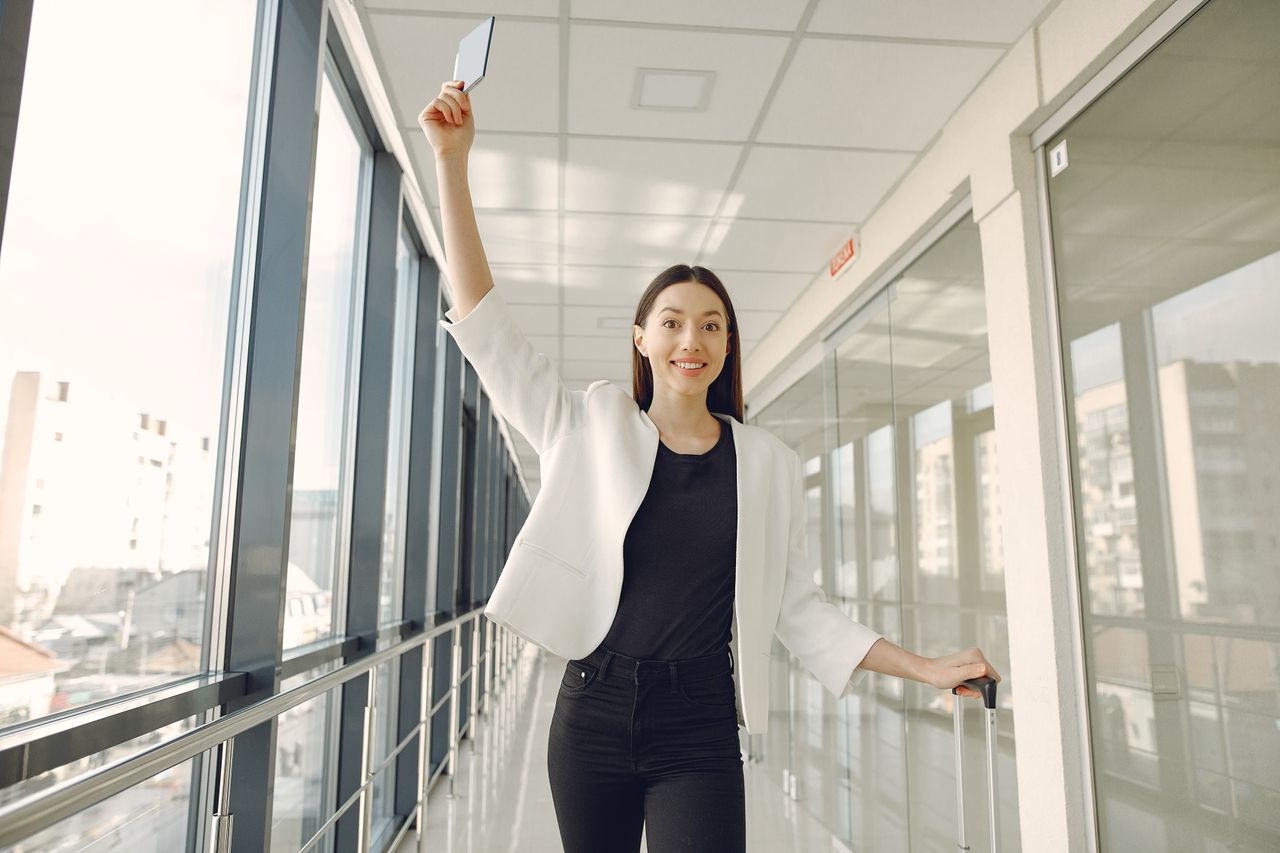 Positive ethnic female traveler in trendy clothes holding passport up for showing documents while walking with baggage along corridor of airport