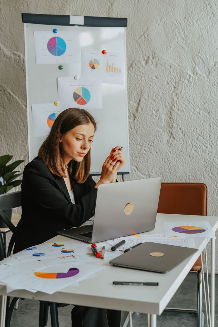 Focused businesswoman working on laptop at desk with charts and graphs in a modern office setting.
