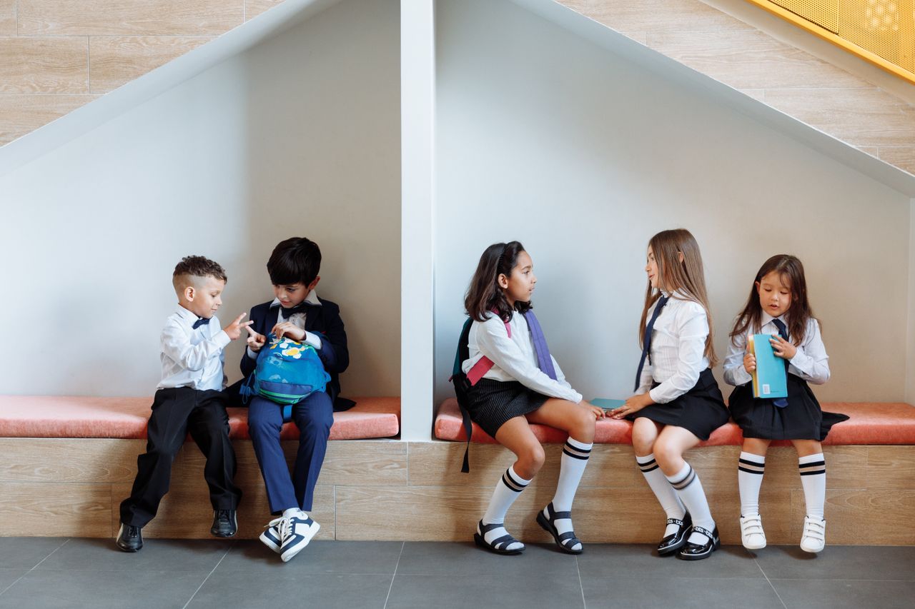 Home Group of boys and girls in school uniforms talking and sitting indoors on a bench.
